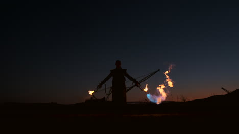 Un-Hombre-Con-Un-Impermeable-Y-Dos-Lanzallamas-Deja-Escapar-Una-Llama-Ardiente-Al-Atardecer-En-La-Arena